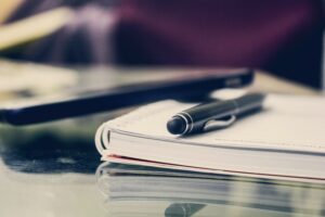 A pen rests on an open notebook placed on a wooden desk, ready for writing or note-taking.